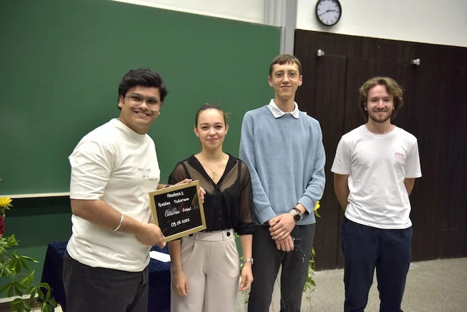 Color photo of four people smiling at the camera, one person holding a prize certificate