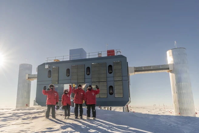 Color photo of 4 people outside in snow standing in front of a large industrial building