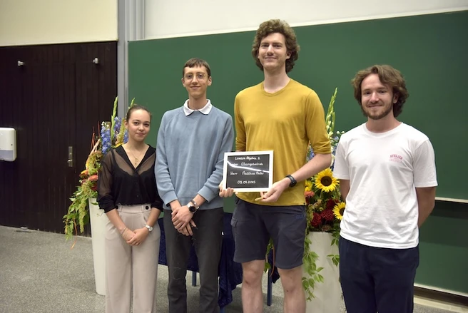 Color photo of four people smiling at the camera, one person holding a prize certificate
