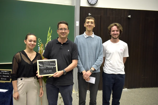 Color photo of four people smiling at the camera, one person holding a prize certificate