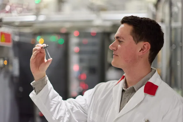 Color photo of a researcher side profile looking left in a lab coat holding a small sample container