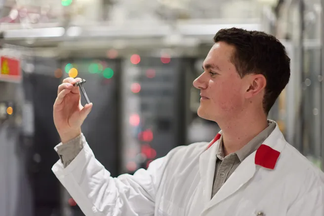 Color photo of a researcher side profile looking left in a lab coat holding a small sample container