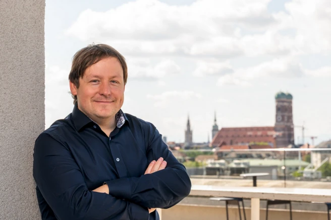 Color photo of a smiling person with crossed arms, a city scene in the background