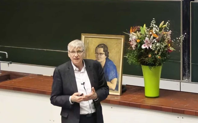Color photo of a person lecturing in a classroom with a portrait and flowers behind them