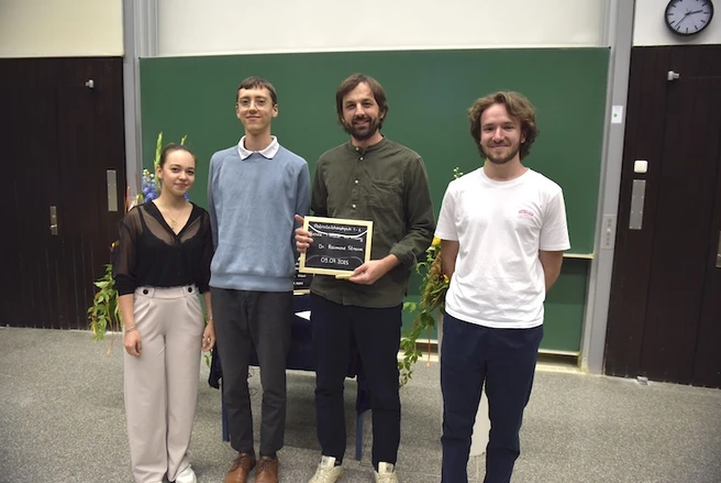 Color photo of four people smiling at the camera, one person holding a prize certificate